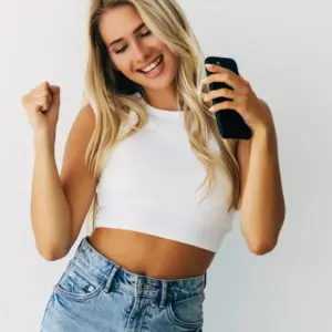 Mujer joven alegre, bailando con móvil en la mañovestida con vaqueros y top de tirantes blanco.